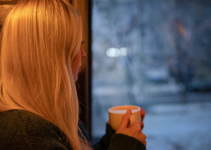 Femme contemplant l'horizon par la fenêtre, une tasse chaude entre les mains, dans une ambiance calme et apaisante.