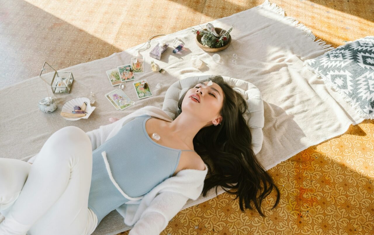 Femme allongée au sol, les yeux fermés, entourée de petits objets bien-être, dans une posture de relaxation ou de méditation.
